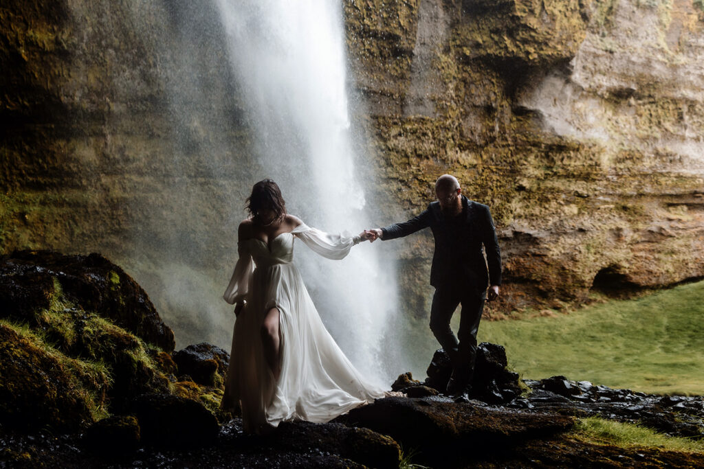 Jade and Bradley holding hands and walking under a waterfall on their elopement day in Iceland